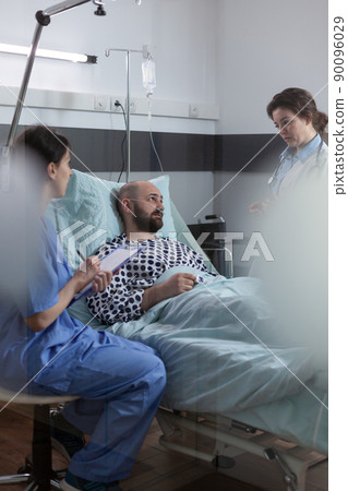Nurse sitting at patient bedside writing prescription medication for pain relief using clipboard. Middle aged man recovering after surgical intervention talks about treatment with doctor. 90096029