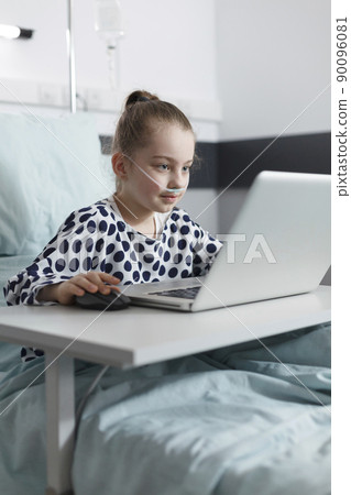 Smiling sick little girl playing games on computer laptop while in pediatric clinic recovery ward room. appy ill little child enjoying video games on laptop while resting in hospital bed. 90096081