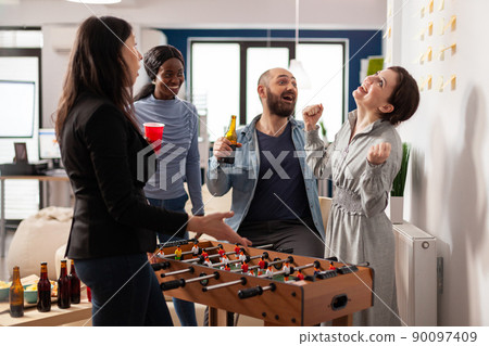 Cheerful woman winning foosball game competition at office party after work, having fun with coworkers. Businesspeople playing soccer match at table, eating snacks and drinking beer. Cheerful woman winning foosball game competition at office party after work, having fun with coworkers. Businesspeople playing soccer match at table, eating snacks and drinking beer. 90097409