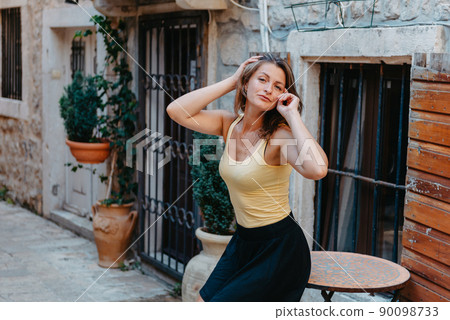 Girl Tourist Walking Through Ancient Narrow Street On A Beautiful Summer Day In MEDITERRANEAN MEDIEVAL CITY, OLD TOWN BUDVA, MONTENEGRO. Young Beautiful Cheerful Woman Walking On Old Street At Girl Tourist Walking Through Ancient Narrow Street On A Beautiful Summer Day In MEDITERRANEAN MEDIEVAL CITY, OLD TOWN BUDVA, MONTENEGRO. Young Beautiful Cheerful Woman Walking On Old Street At 90098733
