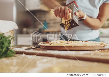 Women's hands grate cheese on a pizza lying on the table in the background of the kitchen Women's hands grate cheese on a pizza lying on the table in the background of the kitchen 90098903