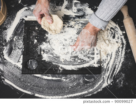two female hands knead the dough from white wheat flour on a black table, top view two female hands knead the dough from white wheat flour on a black table, top view 90099027