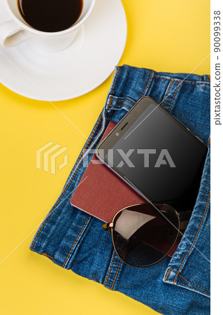 Mobile phone, glasses and passport peek out of the pocket of jeans, next to a cup of coffee 90099338