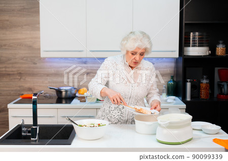 Grandma in the kitchen spreads out steamed salmon and salad on plates Grandma in the kitchen spreads out steamed salmon and salad on plates 90099339