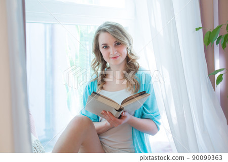 Caucasian teenage girl with a book in the morning sitting on the windowsill against the background of the window 90099363