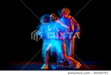 Excited man and woman dancing hip-hop in sportive style clothes on dark background at dance hall in mixed neon light. Youth culture, hip-hop, movement 90099765