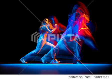 Dynamic portrait of stylish man and girl dancing hip-hop in sportive style clothes on dark background at dance hall in mixed neon light. Youth culture, breakdance, movement 90099835