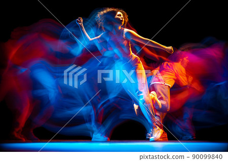 Dynamic portrait of stylish man and girl dancing hip-hop in sportive style clothes on dark background at dance hall in mixed neon light. Youth culture, breakdance, movement Dynamic portrait of stylish man and girl dancing hip-hop in sportive style clothes on dark background at dance hall in mixed neon light. Youth culture, breakdance, movement 90099840