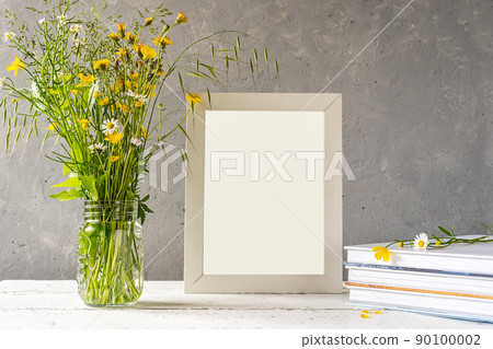 Mock up creation. White frame on white wooden table with grey concrete background, books, notebook and wild flowers Mock up creation. White frame on white wooden table with grey concrete background, books, notebook and wild flowers 90100002