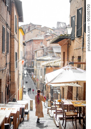 Sylish woman walks on narrow and cozy street in old town of Siena city 90100700