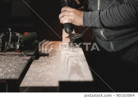 A man works with a drill in his workshop. Carpenter drills with a hand power tool in a dark room with directional light A man works with a drill in his workshop. Carpenter drills with a hand power tool in a dark room with directional light 90103064