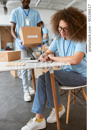 Young team of volunteers in blue uniform working together. Woman making notes and guy carrying cardboard box while working on donation project indoors 90106617