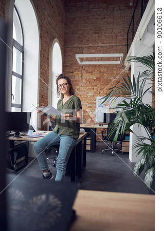 Joyful woman, interior designer or architect wearing glasses smiling at camera while working on a blueprint for new project, leaning on the table in office 90106618