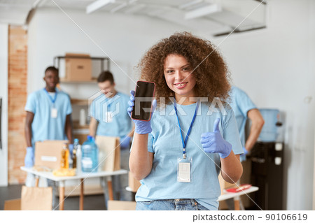 Young woman, volunteer in blue uniform holding, showing smartphone to camera and smiling while standing indoors. Team sorting, packing items in the background 90106619