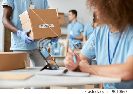 Young female volunteer in blue uniform making notes while working on donation project. Team sorting, packing items in cardboard box in the background 90106623