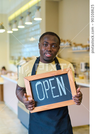 Portrait of African young waiter in uniform holding open sign and looking at camera, he opening new coffee shop Portrait of African young waiter in uniform holding open sign and looking at camera, he opening new coffee shop 90108625