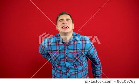 A young man has a backache and is holding it in his hands. Shooting on a red background. A young man has a backache and is holding it in his hands. Shooting on a red background. 90109762