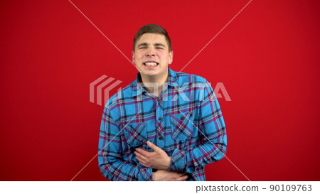 A young man has a stomach ache and he holds it in his hands. Shooting on a red background. A young man has a stomach ache and he holds it in his hands. Shooting on a red background. 90109763