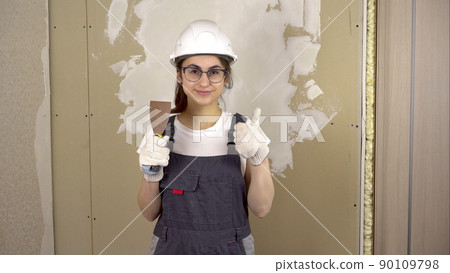 A builder with a spatula in his hand looks at the camera and shows the class and smiles. A woman in special clothes and a helmet makes repairs. 90109798