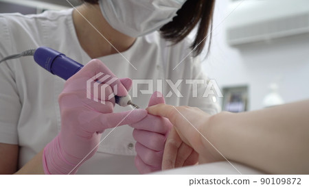 A professional manicurist using a drill tool cuts off old nails close-up. Nail care in a manicure salon. A professional manicurist using a drill tool cuts off old nails close-up. Nail care in a manicure salon. 90109872