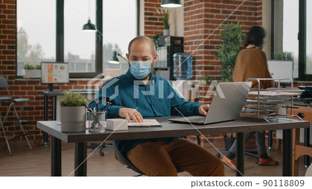 Business man doing paperwork and looking at rate charts while working with laptop. Employee wearing face mask and analyzing data analysis on files for project planning and strategy. 90118809