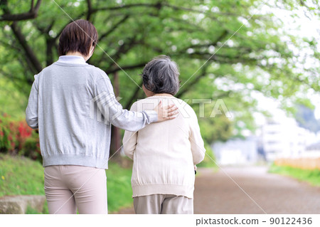 Senior woman taking a walk in the park and a caregiver rehabilitating behind Senior woman taking a walk in the park and a caregiver rehabilitating behind 90122436