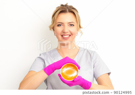 Portrait of a female caucasian beautician holding a jar of sugar paste for sugaring wearing pink gloves on white background Portrait of a female caucasian beautician holding a jar of sugar paste for sugaring wearing pink gloves on white background 90124995