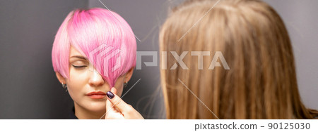 Hairdresser with hands is checking out and fixing the short pink hairstyle of the young white woman in a hair salon 90125030