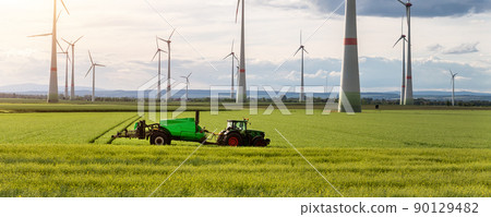 Scenic landscape view big modern tractor machine with sprayer equipment spraying fertilizer in rapeseed agricultural field against windfarm power windmill. Arable land cultivation sustainable energy Scenic landscape view big modern tractor machine with sprayer equipment spraying fertilizer in rapeseed agricultural field against windfarm power windmill. Arable land cultivation sustainable energy 90129482