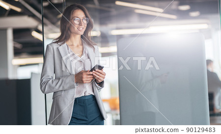 Businesswoman wearing glasses with mobile phone in hand standing in modern office 90129483