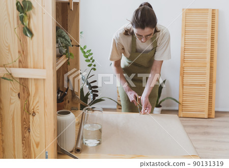 Woman seller in a green aprone cuts craft paper with scissors. Flower and gift shop Woman seller in a green aprone cuts craft paper with scissors. Flower and gift shop 90131319