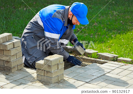 Bricklayer in work clothes sits on sidewalk and lays out paving slabs. Sight of working man in open 90132917