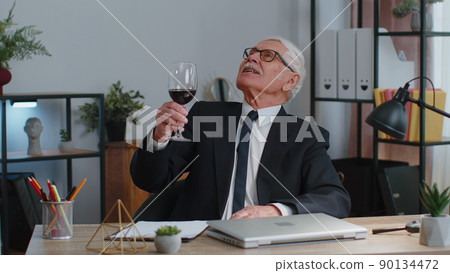Senior man freelancer drinking wine after working on laptop computer pc sitting at desk at office 90134472