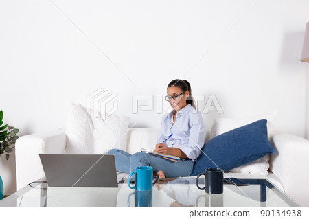 Young adult woman taking notes with her laptop on the couch in the living room at home. With copy space. Young adult woman taking notes with her laptop on the couch in the living room at home. With copy space. 90134938