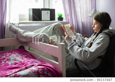 A teenage girl reads her school reading while sitting comfortably by the window in her room. A teenage girl reads her school reading while sitting comfortably by the window in her room. 90137887