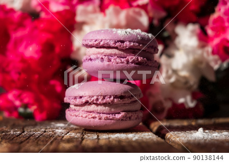 Purple creamy macarons with sugar powder on the wooden board, pink bouquet of flowers in the background Purple creamy macarons with sugar powder on the wooden board, pink bouquet of flowers in the background 90138144