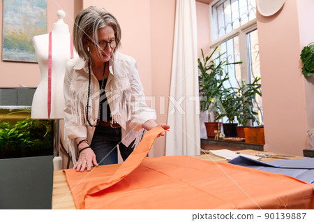 Charming mature female fashion designer tailor spreading new type of luxurious tissue on a wooden table before putting a sewing pattern and cutting for creating new garment in tailoring atelier 90139887