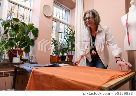Stylish mature Caucasian woman seamstress spreading new fabric in workshop while creating new collection of clothes from luxurious tissue Stylish mature Caucasian woman seamstress spreading new fabric in workshop while creating new collection of clothes from luxurious tissue 90139888