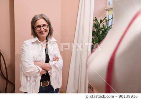 Elegant mature European woman dressmaker, fashion designer, smiling, standing with her arms crossed next to a mannequin in her own fashion atelier Elegant mature European woman dressmaker, fashion designer, smiling, standing with her arms crossed next to a mannequin in her own fashion atelier 90139916