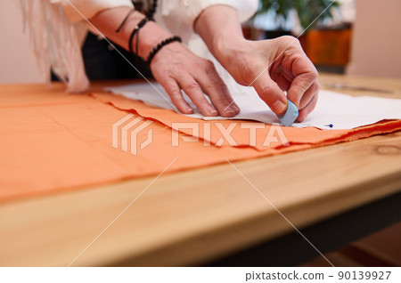 Tailor at work. Close-up of a dressmaker's hands transferring sewing patterns for clothes on orange colored satin fabric in fashion design atelier 90139927
