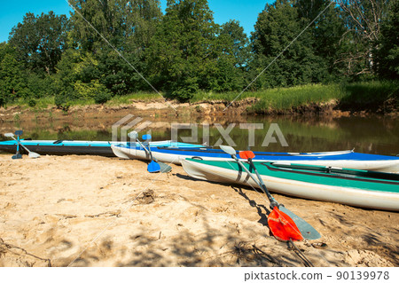 Tourist canoes with paddles stand on the river coast in summer on a water hike. Rafting on inflatable and frame double and triple kayak boats, family trip, extreme adventure in summer Tourist canoes with paddles stand on the river coast in summer on a water hike. Rafting on inflatable and frame double and triple kayak boats, family trip, extreme adventure in summer 90139978