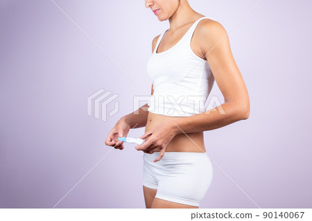 Young caucasian woman in white T-shirt and shorts, holding pregnancy test on neutral background. Woman and pregnancy test concept. Young caucasian woman in white T-shirt and shorts, holding pregnancy test on neutral background. Woman and pregnancy test concept. 90140067