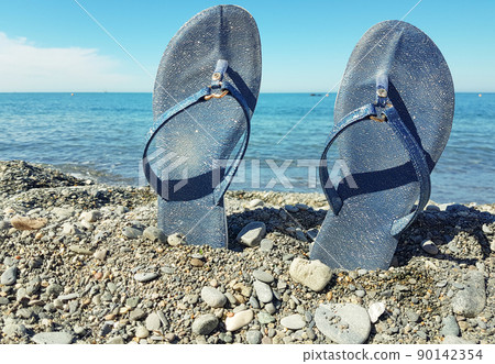 Blue women's beach shoes, standing in the pebbles against the background of the sea and the blue sky. Close-up, a place for text, the concept of a beach holiday by the sea Blue women's beach shoes, standing in the pebbles against the background of the sea and the blue sky. Close-up, a place for text, the concept of a beach holiday by the sea 90142354