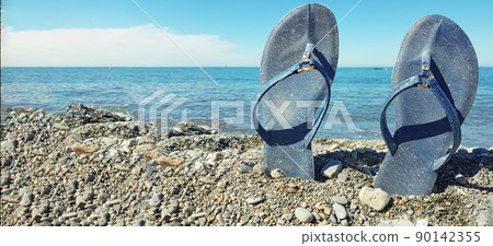 Blue women's beach shoes, standing in the pebbles against the background of the sea and the blue sky. Close-up, place for text, concept of a beach holiday by the sea, banner 90142355