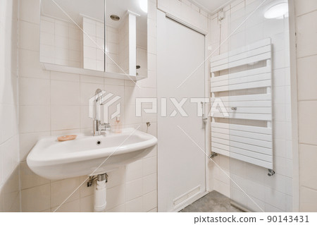 Cupboard with sink and mirror attached to tiled wall near door and drying rack in modern restroom Cupboard with sink and mirror attached to tiled wall near door and drying rack in modern restroom 90143431