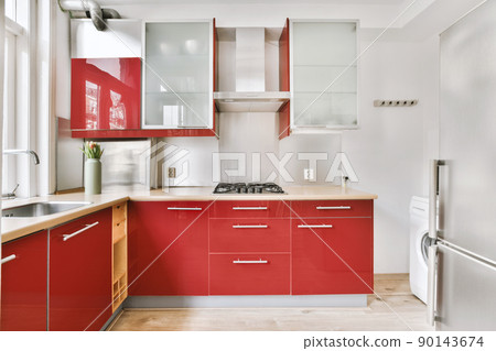 Small corner kitchen with sink under window and red furniture with transparent cupboards with stove, fridge and washing machine in urban apartment 90143674