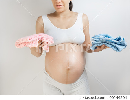 A woman in anticipation of a girl and a boy twins prepares gifts for their birthday. Baby Clothing in mother hands. Pregnant woman holding boy and girl clothes. Expecting newborn baby and guessing 90144204
