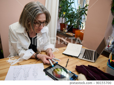 Mature woman fashion designer working on color wheel on a digital tablet, holding watercolor markers while sketching clothes in a tailoring atelier 90155700
