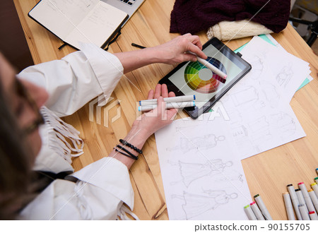 Mature European confident woman, senior fashion designer choosing color palette. Matches color markers to the color wheel while coloring clothes sketches of new collection 90155705