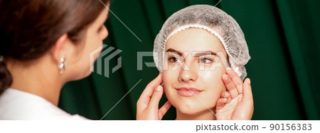 Plastic surgery and beauty concept. Professional cosmetician examining and touching the face of the beautiful young caucasian smiling woman in a clinic of esthetic cosmetology Plastic surgery and beauty concept. Professional cosmetician examining and touching the face of the beautiful young caucasian smiling woman in a clinic of esthetic cosmetology 90156383